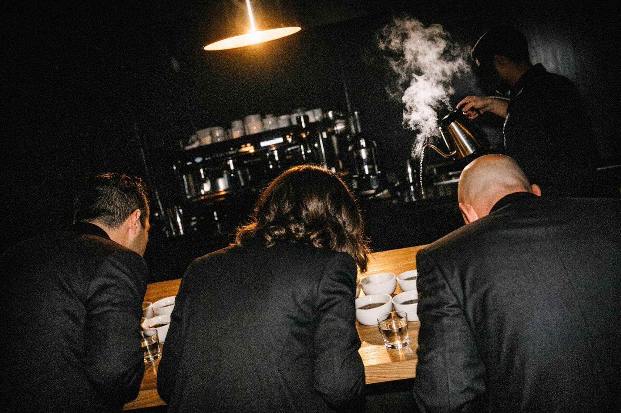 Three people in dark clothes seen from behind leaning over coffee cupping bowls at a private after-hours tasting, steam lit by flash