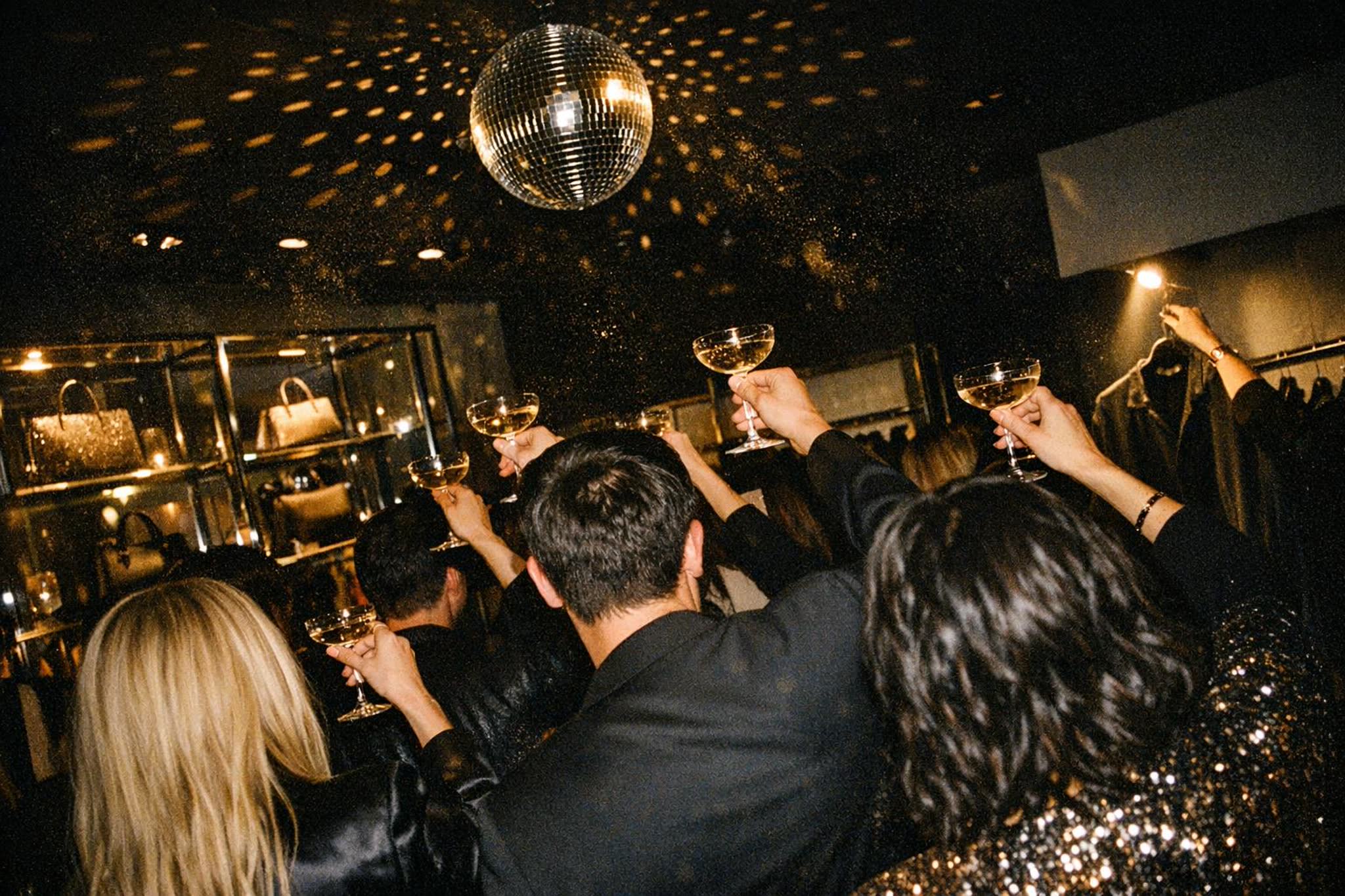 Chandelier-lit salon full of guests in evening wear; one holds a brocade jacket up on its hanger over a champagne table