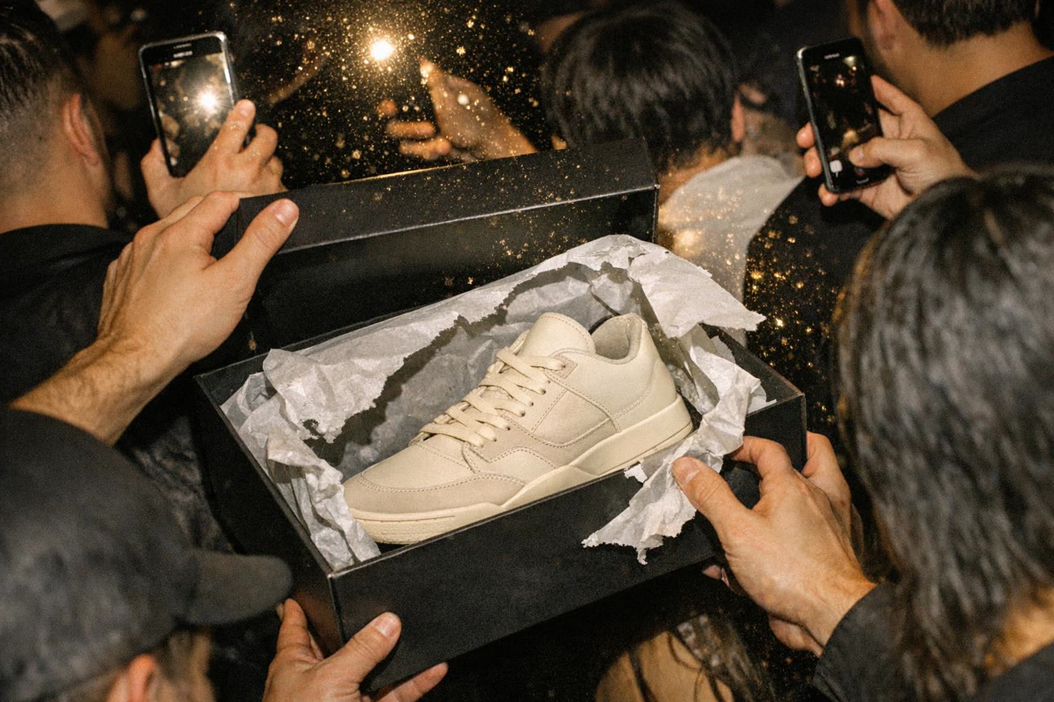 Hands with gold rings lift tissue paper from a black box while a crowd films the reveal on their phones
