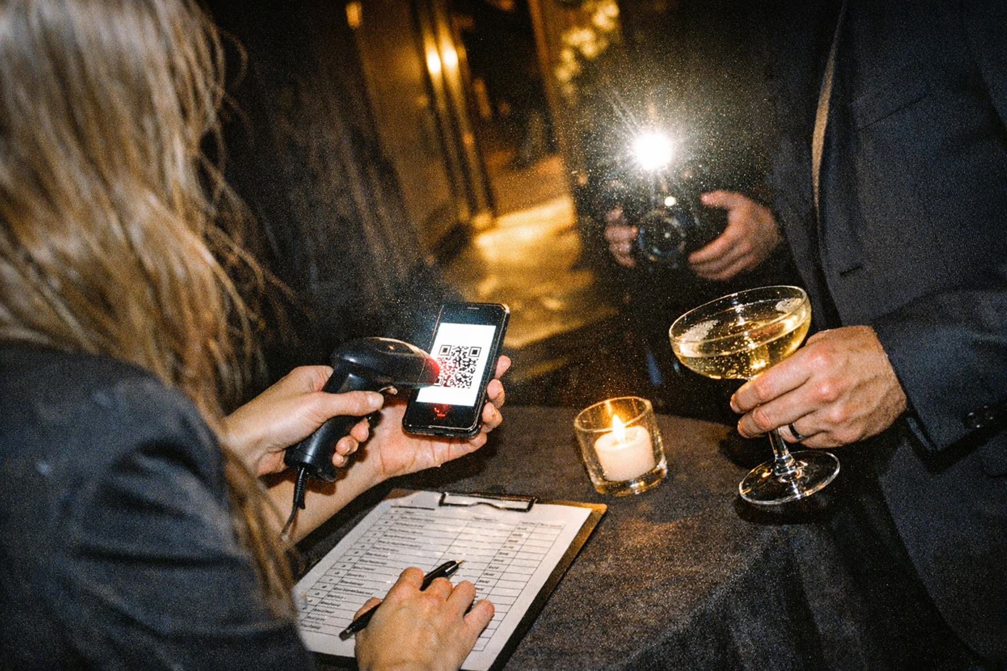 A host scans the QR pass on a guest's phone at the door of a night event while their plus-one waits beside them