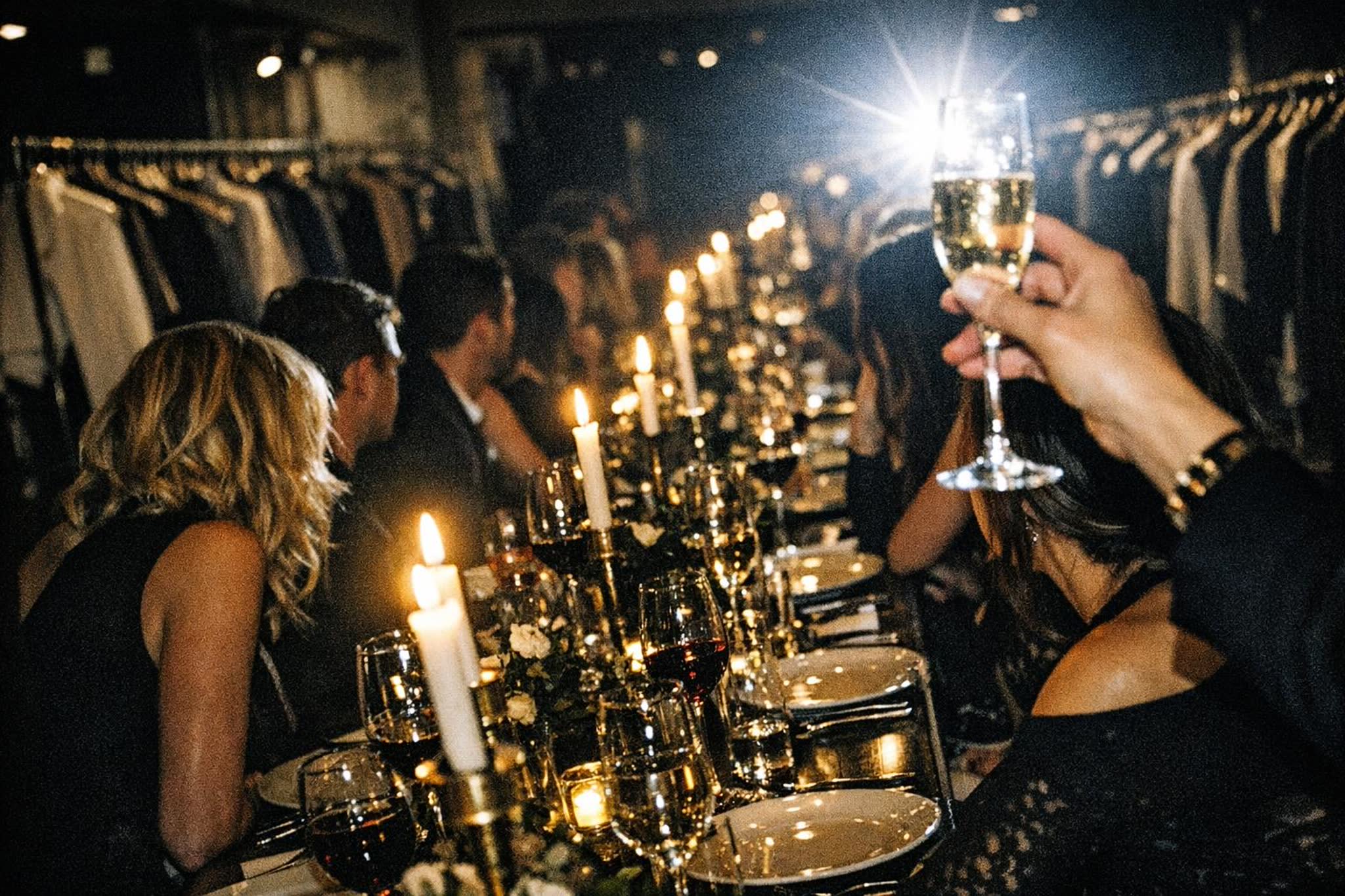 A long dinner table set between the clothing racks of a store after hours, guests seated under warm low light