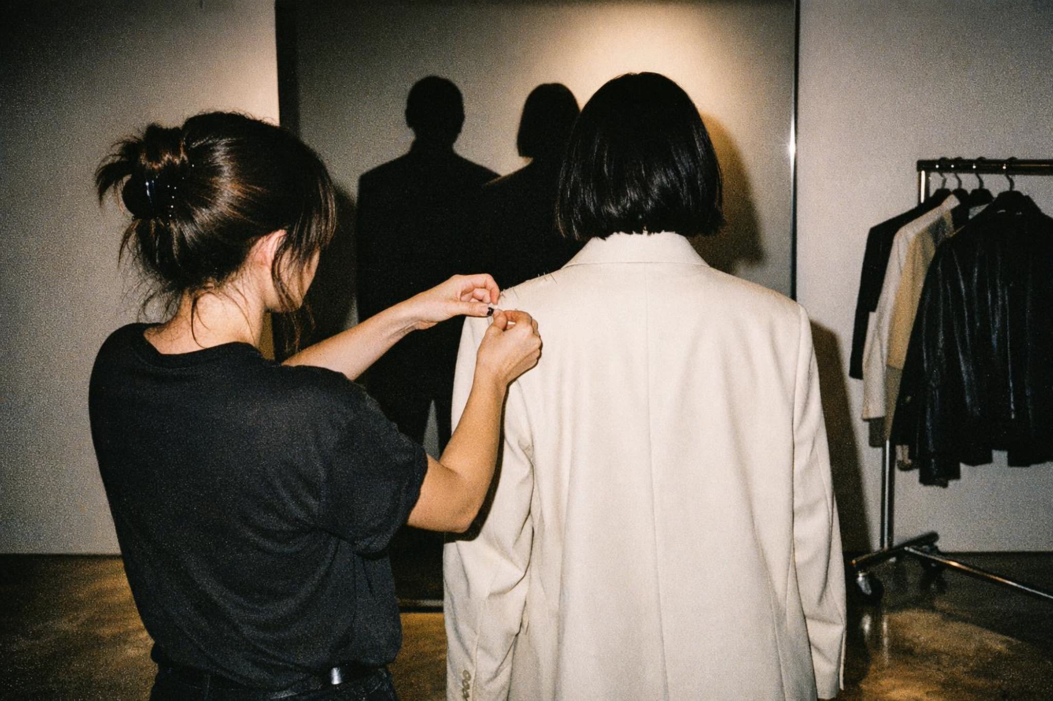A stylist seen from behind pinning the shoulder of an oversized cream blazer on a client facing a mirror in a modern boutique