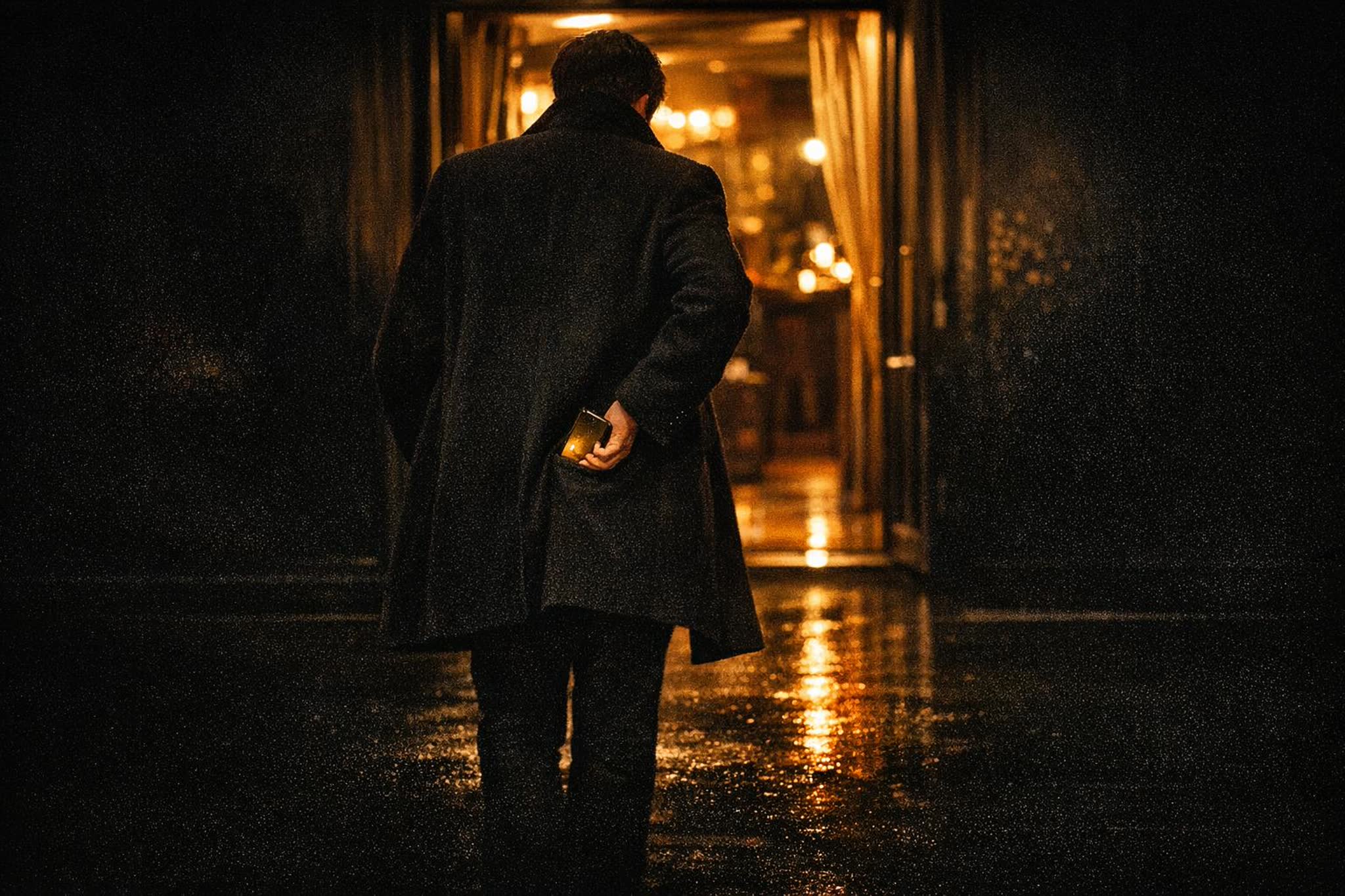 A guest in a long coat seen from behind slipping a glowing phone into his pocket while walking into a warmly lit doorway at night